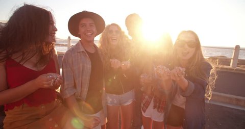Group of teenager hipster friends celebrating by blowing colorful confetti from hands with sunset sun flare