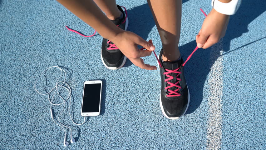 Closeup of feet of female runner getting ready tying running shoes with smartwatch, earphones and phone for music motivation for cardio workout training on athletic track in outdoor gym.