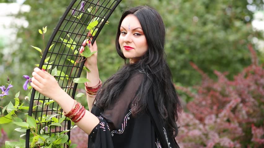 Brunette woman in black dress standing near lattice arch in park and posing.
