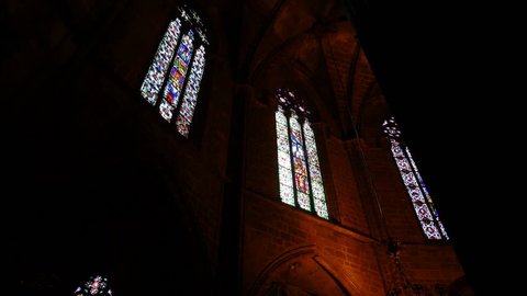 Colorful stained-glass windows at Gothic Cathedral, POV camera move inside, Ambulatory of Saint Eulalia basilica. Low angle shot, look upward to decorated lanced windows, glowing at dark interior.