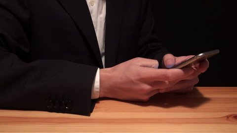 Businessman in a suit typing in smartphone, wood table, black background. Man dressed in dark jacket and light shirt write text messages or email on your smartphone-Adrian