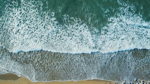 Aerial drone footage of serene sea waves reaching shore. Slow lockdown shot of textures being formed by white sea foam. As the waves calm the patterns change. Filmed from an overhead perspective.