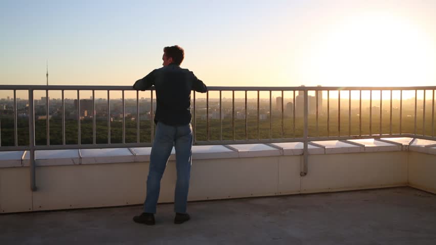 Man walking on roof back and forth along fence at sunset.
