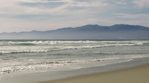 View along the coast line, slow motion looking across Venice Beach towards Santa Monica with rolling breakers and boats on the Pacific Ocean.  Santa Monica Mountains further in the background.