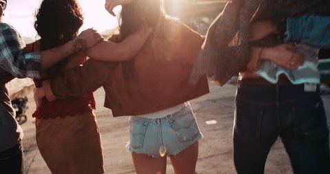 Back view of a group of teen hipster friends enjoying a walk on the pier hugging each other with sunset sun flare