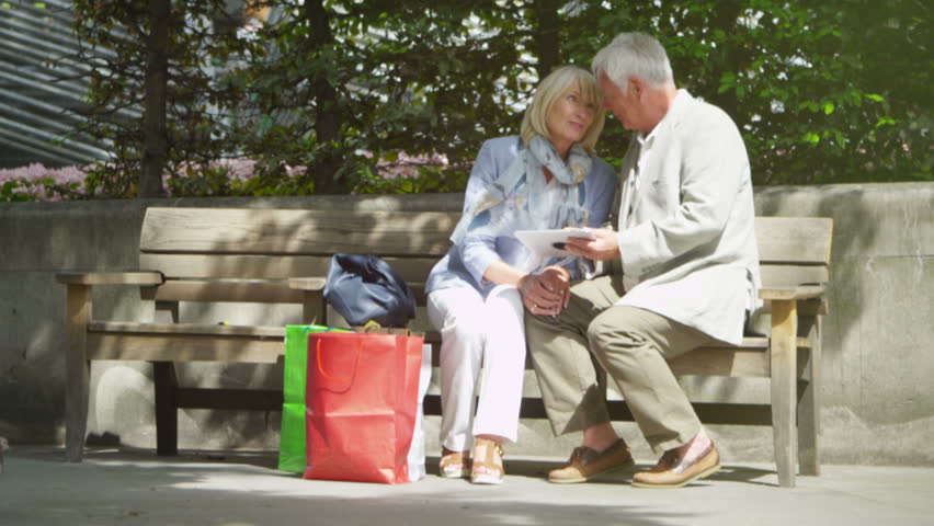 4K Happy mature couple with shopping bags, looking at computer tablet outdoors