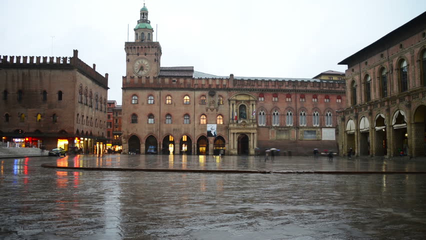 Heavy rain in Bologna, Italy with people walking by the Piazza Maggiore holding umbrellas. Central square in the city during the evening in spring
