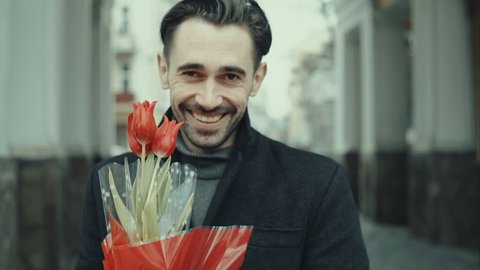 Happy smiling forty years old caucasian man with baguette and flower bouquet standing on the street. City buildings as background. RAW video rcord.