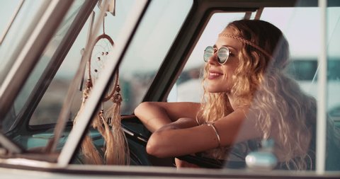 Blonde boho Hipster girl sitting and smiling in front drivers seat of retro van parked at sandy beach during sunset, resting from Road Trip