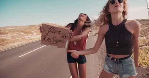 Two friends on a road trip; Blonde caucasian hipster girl and african american boho woman hitchhiking on roadside of highway with thumb out and holding cardboard sign viva Las Vegas