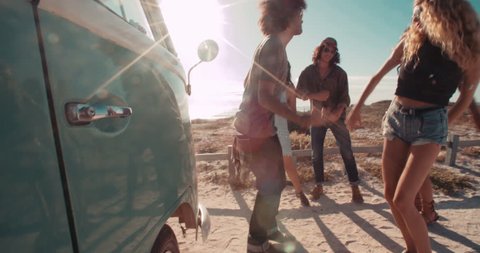 A group of multi-ethnic hipster road trip friends laughing and talking while dancing next to a parked retro van next to a sandy beach during sunset