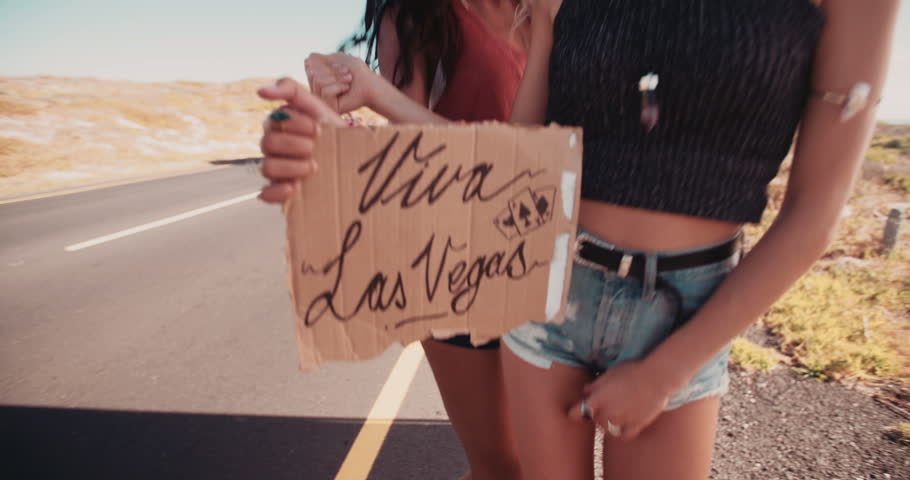 Two friends on a road trip; Blonde caucasian hipster girl and african american boho woman hitchhiking on roadside of highway with thumb out and holding cardboard sign viva Las Vegas