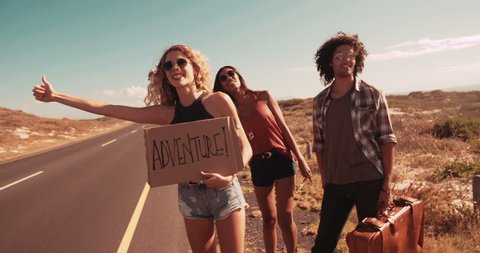 Hipster friends in the daytime stand on side of two lane highway hitchhiking while holding cardboard sign with the word Adventure written on it and sticking out thumb