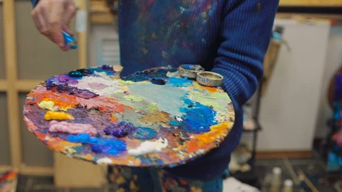 artist brush mix color oil painting on palette is holding in his hand closeup. In workshop
