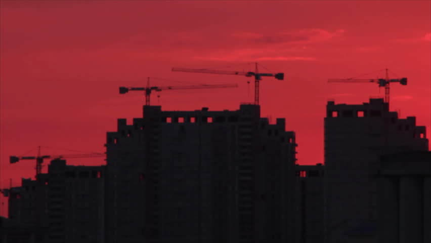 Silhouettes cranes and construction buildings at backdrop darkening red dusk sky