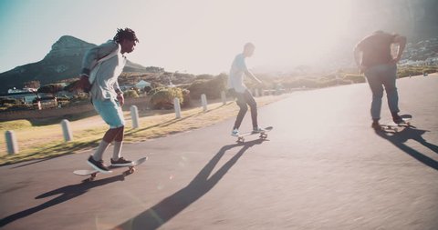 Multi-Ethnic group of skater friends skateboarding down road at seaside together during sunset