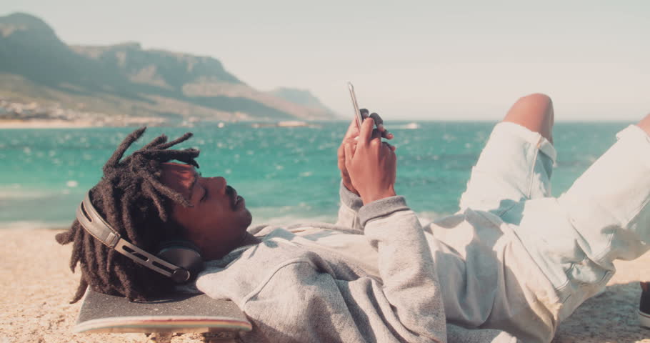 African American skater lying down , listing to music from his smartphone and resting his head on his skateboard on the rocks of a seaside during sunset