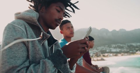 African American looks at smartphone and sits outside while mixed race friend sits beside him at the seaside during sunset