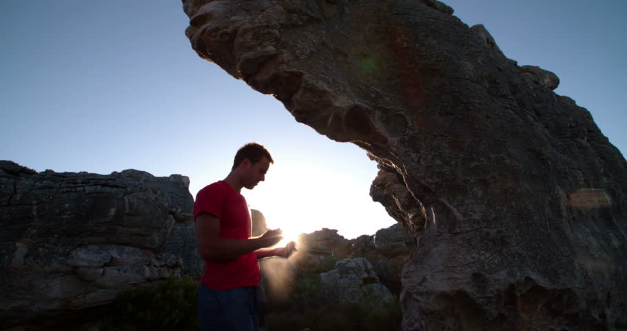 Man backlit from sunlight agains rock nature background. In preparation for bouldering and climbing he rubs chalk onto his hands. He is focussed on the ascent.