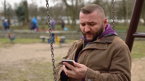 Sad man writing a message and sitting at the swing and swinging 
