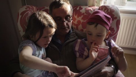 A grandfather is reading a story book to his granddaughter and grandson. The grandson is dressed up as a princess from the story in the gender blender style. Shot in a modern British house.