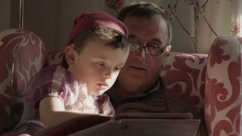 A grandfather is reading a story book to his grandson. The grandson is dressed up as a princess from the story in the gender blender style. Shot in a modern British house.
