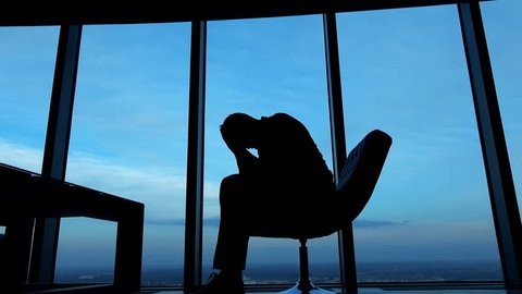 Silhouette of sad, unhappy businessman sitting on armchair by the window, against the sky, 4K
