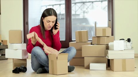 Young woman looks disappointed after shopping on line and complaining by phone with laptop and stack of boxes on the floor

