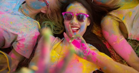 Multi-Ethnic group covered in Holi powder lay in grass laughing and looking up and gesturing peace sign to the camera in a park at a Holi festival in the summer