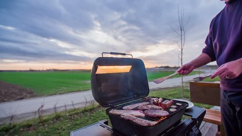 A man is cooking meat on the barbecue grill. The sunset behind him is amazing. He is preparing some dinner for him and his wife.