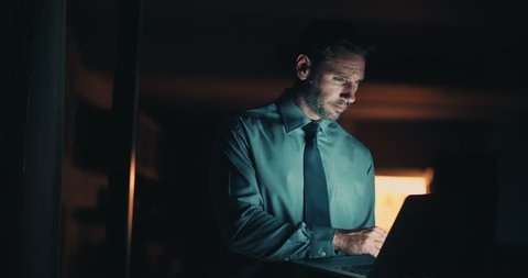 Serious man in blue shirt and blue tie working on laptop at night time in office