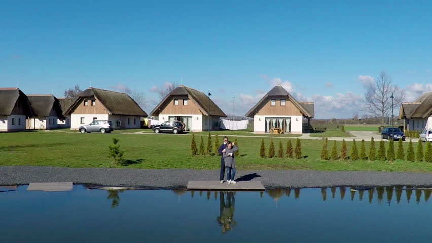 We can see beautiful blue water of the pond and the holiday homes in the background. A senior couple is standing on the bank.