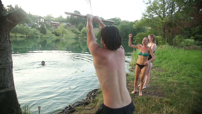 Handsome young man jumping off rope swing into river while happy friends cheer for him, in slow motion