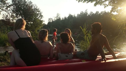 Back view of the group of five young friends resting in nature after riding a canoe, talking and laughing by the river at sunset : vidéo de stock