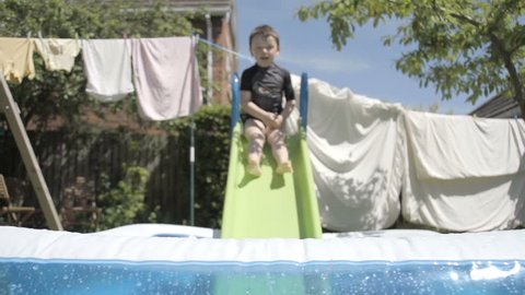 Little Boy Having Fun In A Paddling Pool In Slow Motion. A little boy is enjoying a hot summers day playing in the back garden with a slide and a paddling pool