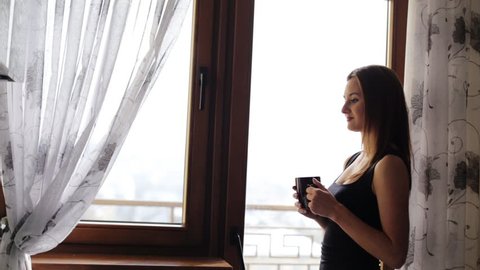 Attractive young lady having a rest and drinking a cup of tea or coffee in front of the window at home.