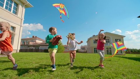 Four kids are running on green grass: two girls flying kites, boys holding wooden toys