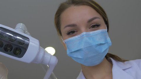 Young Female Friendly Dentist in Mask is Smiling, Holding a Mouth Mirror and Hook, Turns the Light of Dental Lamp On, Lights Into a Patient's Mouth, Standing Upon a Patient, Looking at Camera,trying