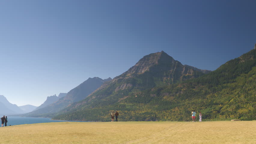 Tourists walking on grass overlook near Waterton Lake and Prince of Wales Hotel, Waterton Lakes National Park, Alberta, Canada. 4K.