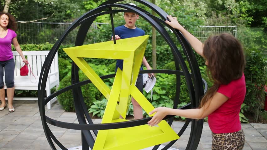 Girl and boy rotating sphere prisms inside, and their mother sitting near on park bench.