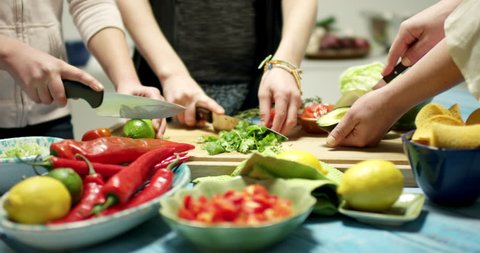 Several hands preparing for a taco tortilla dinner in a kitchen. Cutting fresh vegetables on a wooden bench in a modern kitchen with shallow focus and blurred background. Strong and vivid colors.