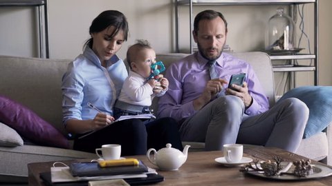 Business family with small baby working with smartphone and documents on sofa
