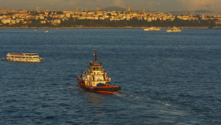 An orange coastal safety boat patrols the waters of the Bosporus Strait at Istanbul, Turkey at sunset as a ferry boat passes.  Tree-line shore with city, mountains, clouds and the Parliament building