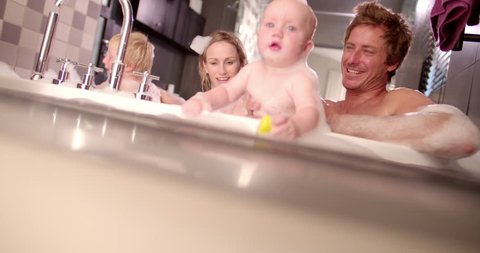 Family of four: mother, father, baby girl, little son taking a big bubble bath together filled with soap suds and smiling