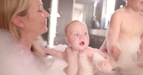 Family of mom, dad and infant daughter taking a bubble bath together and playing in the soap suds in a sunlight lit bathroom