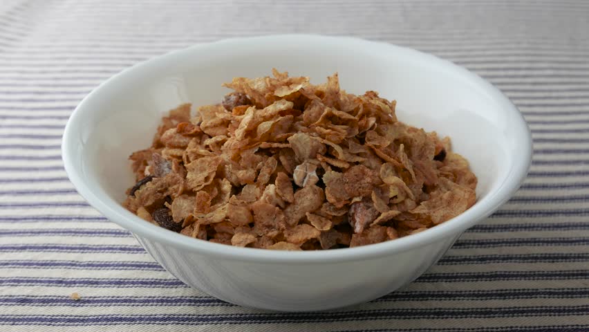 A bowl of pecans, raisins and dates breakfast cereal having skim milk sloppily poured onto the cereal upon a striped table cloth.