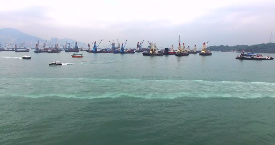 Aerial view of a lot of different ships at anchorage and a few holiday cruisers floating in the bay of Hong Kong, China. Hills are seen on the background across the bay.		