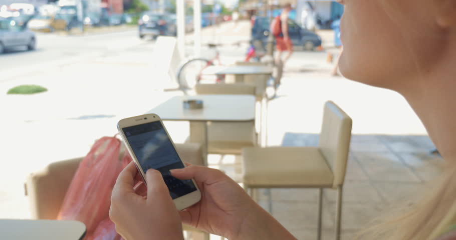 Young blond woman in sunglasses getting in touch with sms.  She sitting in city street cafe on bright summer day, texting and sending message