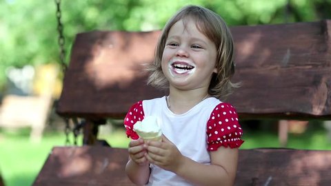 Little girl sits on the swing bench eats ice cream smiles and laughs. Very funny girl