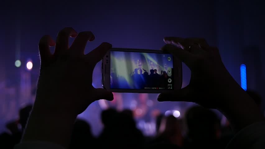 KHERSON, UKRAINE - MAR 26, 2016 - Free public music concert - Fan spectator shooting video via smartphone camera at concert flashing spotlight lumiere | Shutterstock HD Video #15623020